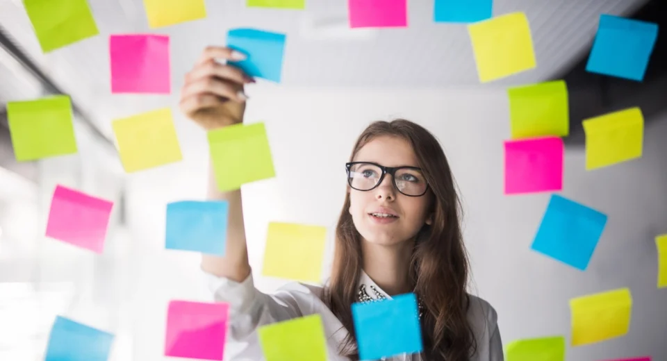 Mulher analisando post-its em parede de vidro, representando a definição do perfil ideal de cliente.