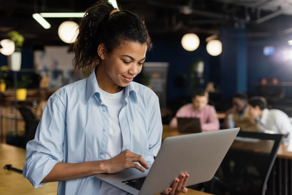 Mulher sorridente trabalhando com laptop no escritório, simbolizando os primeiros passos para empreender do zero.