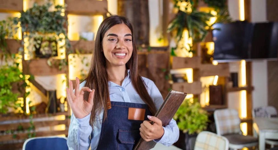 Garçonete sorridente segurando um menu em restaurante, ilustrando um exemplo de serviço com baixo custo para empreender.
