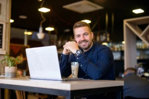 Homem sorridente trabalhando em seu laptop em um café, simbolizando o início de um negócio com poucos recursos.