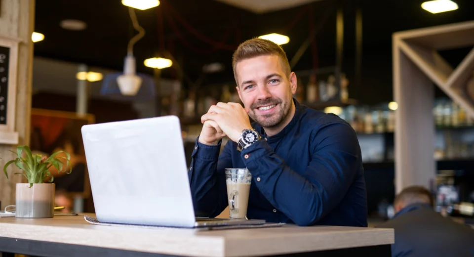Homem sorridente trabalhando em seu laptop em um café, simbolizando o início de um negócio com poucos recursos.